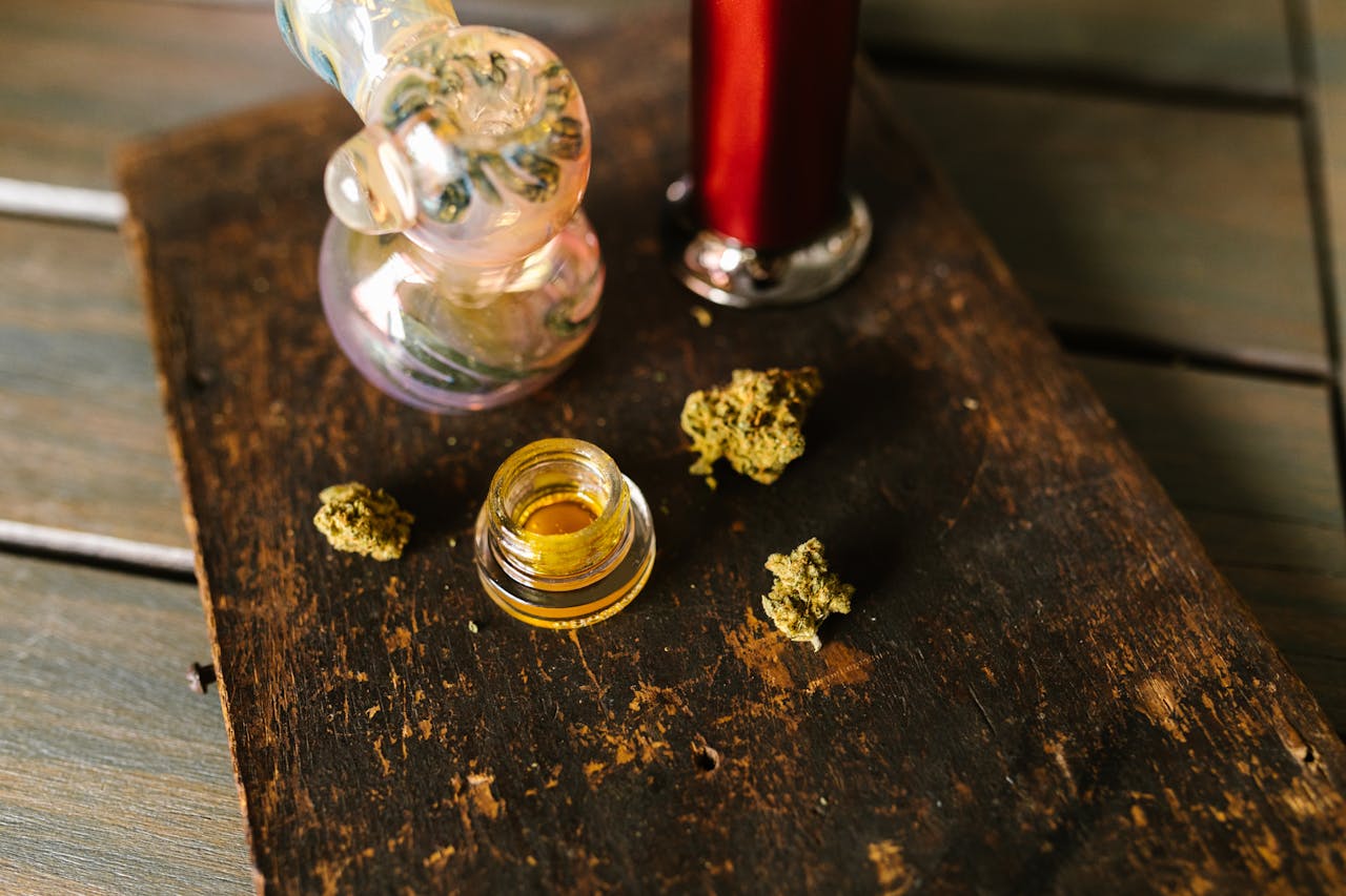 Close-up of cannabis buds, glass pipe, and wax on wooden surface.